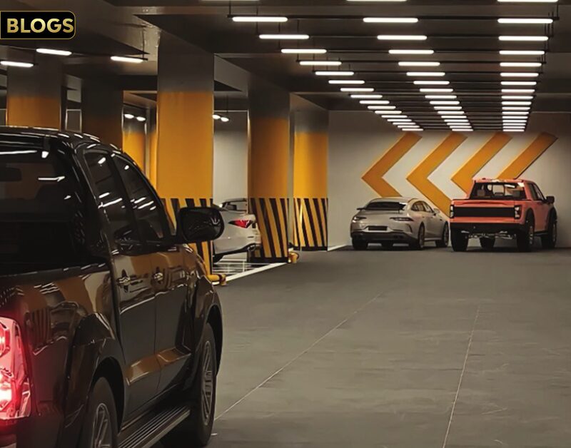 MGI Paradise: Your Premier Living Experience: The image shows a modern, well-lit underground parking garage with multiple vehicles parked. The garage features bright yellow and black striped columns and directional arrows on the walls, guiding the flow of traffic. The clean and organized space suggests a high-end residential or commercial building, emphasizing convenience and luxury.